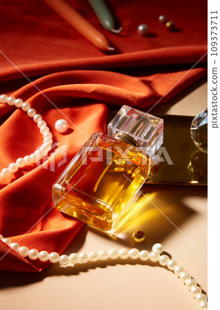 A bottle of perfume without label is displayed on a table with a string of pearls, candles and red silk. Mockup for perfume advertisement with blank label. 109373711