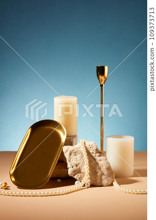 Front view of two white candles placed next to a stone slab, string of pearls and props on a pastel brown table with a blue background. Scene for advertising. 109373713