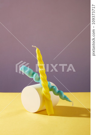 Two twisted candles in two colors: yellow and blue are placed next to a white podium on a yellow table with a purple background. Creative space for decoration. 109373717