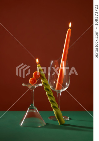 Two burning candles are placed next to a cocktail glass and a wine glass on a deep green and brown background. Mysterious space for advertising and some other purposes. 109373723
