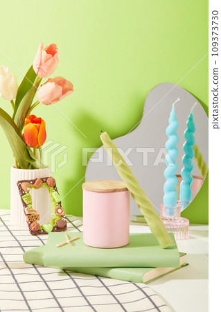 Tulips are placed in a porcelain vase. A jar of scented candles is displayed on the table with two notebooks. Colored candles and a mirror are placed on the table. Mockups for ads. 109373730