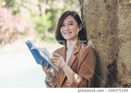 Happy asian woman reading book outdoor in green park nature. Reading book magazine relaxation woman. Leisure portrait woman smile sitting in green park read e-book diary planner with happy face Happy asian woman reading book outdoor in green park nature. Reading book magazine relaxation woman. Leisure portrait woman smile sitting in green park read e-book diary planner with happy face 109375109