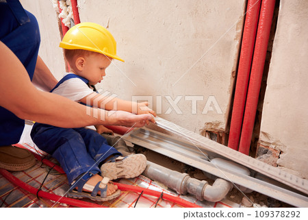 Man and little boy sitting by the wall with pipes and holding material for plumbing installation. Male plumber and child in safety construction helmet working together on home renovation. 109378292