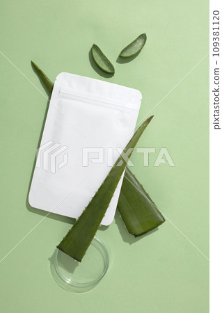 A non-branded mask sheet rests on a fresh aloe vera leaf, beside a clear petri dish, set against a pastel background-a cosmetic brand mockup. Copy space. 109381120