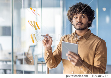 Portrait successful businessman inside office, man serious looking at the camera with a tablet computer in hands, standing near a glass board with colored notes, business strategy planning. Portrait successful businessman inside office, man serious looking at the camera with a tablet computer in hands, standing near a glass board with colored notes, business strategy planning. 109383216