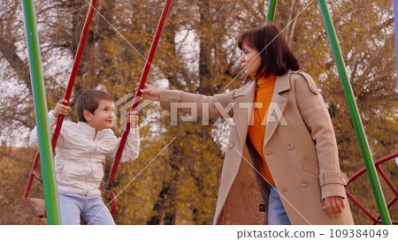 Mother swings boy on swing on park playground. Mother swings preschooler boy on vividly hued swing in park. Mother swings boy in autumn park on brightly colored swing creating memorable family fun Mother swings boy on swing on park playground. Mother swings preschooler boy on vividly hued swing in park. Mother swings boy in autumn park on brightly colored swing creating memorable family fun 109384049