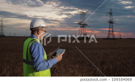 Mature employee with helmet conducts digital expertise of power distributions substation via tablet in sunset field. Engineer with tablet controls power generation substation in sunset field Mature employee with helmet conducts digital expertise of power distributions substation via tablet in sunset field. Engineer with tablet controls power generation substation in sunset field 109384069
