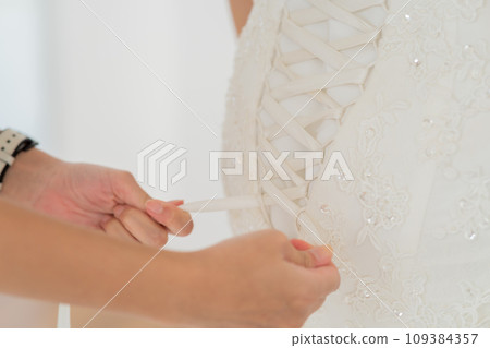 Close-up of hands meticulously tying the lace on a bridal gown, details of wedding day preparations 109384357