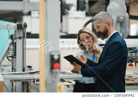 Female engineer and male production manager standing in modern industrial factory, talking about production. Manufacturing facility with robotics and automation. Female engineer and male production manager standing in modern industrial factory, talking about production. Manufacturing facility with robotics and automation. 109384942
