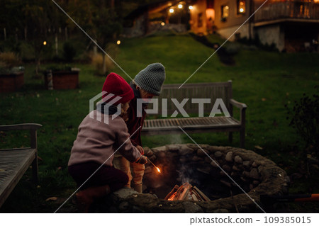 The family spending evening outdoors, lighting fire in a fire pit. Prents and kids spending quality family time together. The family spending evening outdoors, lighting fire in a fire pit. Prents and kids spending quality family time together. 109385015