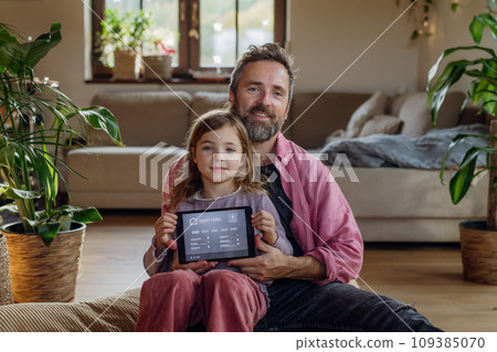 Father and girl holding smart thermostat, adjusting, lowering heating temperature at home. Concept of sustainable, efficient, and smart technology in home heating and thermostats. Father and girl holding smart thermostat, adjusting, lowering heating temperature at home. Concept of sustainable, efficient, and smart technology in home heating and thermostats. 109385070