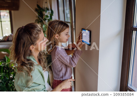 Girl helping mother to adjust, lower heating temperature on thermostat. Concept of sustainable, efficient, and smart technology in home heating and thermostats. Girl helping mother to adjust, lower heating temperature on thermostat. Concept of sustainable, efficient, and smart technology in home heating and thermostats. 109385084
