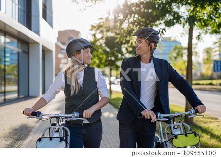 Colleagues commuting through the city, talking and walking by bike on street. Middle-aged city commuters traveling from work by bike after a long workday. Colleagues commuting through the city, talking and walking by bike on street. Middle-aged city commuters traveling from work by bike after a long workday. 109385100