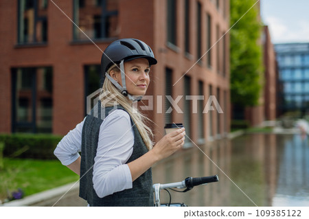 Beautiful middle-aged woman commuting through the city, buying, drinking coffe in front of office. Female city commuter traveling from work by bike after a long workday. 109385122