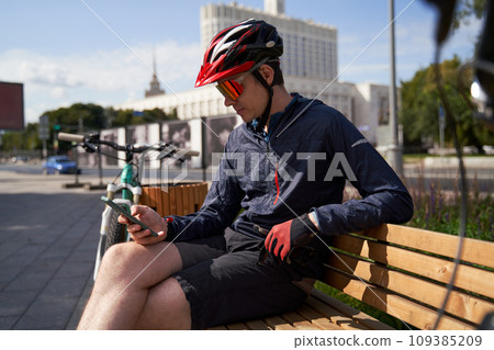 Man in a bicycle helmet sits on a bench and looks at his phone, resting while cycling in the city 109385209