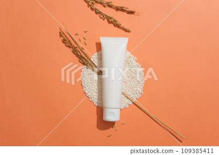 A tube without label placed on a pile of rice and decorated with wheat ears. Orange background. Empty label for cosmetic product mockup 109385411
