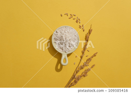 Against yellow background, a ceramic bowl of rice bran is featured with wheat ears. Rice bran in skincare is beloved because it can be used daily Against yellow background, a ceramic bowl of rice bran is featured with wheat ears. Rice bran in skincare is beloved because it can be used daily 109385416