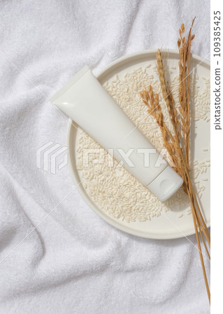 White cosmetic tube without label arranged on a dish in round shaped with lots of rice. Some wheat ears are featured. Rice water is a fairly common ingredient in products 109385425