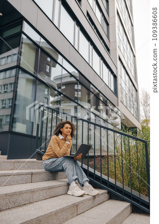 Focused woman manager working laptop sitting on stairs on modern building background Focused woman manager working laptop sitting on stairs on modern building background 109386608