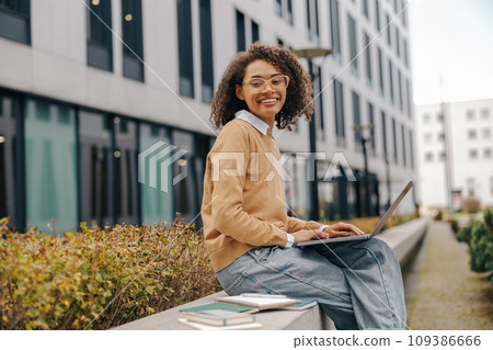 Smiling woman manager in eyeglasses is working on laptop sitting on background of office building Smiling woman manager in eyeglasses is working on laptop sitting on background of office building 109386666