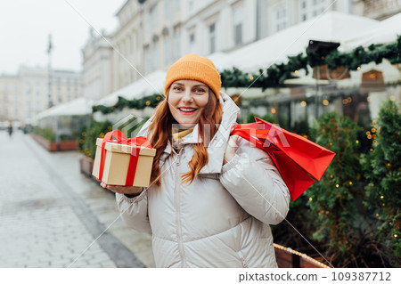 Happy cheerful young Woman holding red shopping paper bags and present in hands, walking at street. Winter holidays sale. Shopping banner, for mockup Happy cheerful young Woman holding red shopping paper bags and present in hands, walking at street. Winter holidays sale. Shopping banner, for mockup 109387712