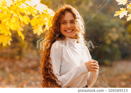 Smiling young woman enjoy the autumn weather in the parkland with the yellow leaves at branches Smiling young woman enjoy the autumn weather in the parkland with the yellow leaves at branches 109389315