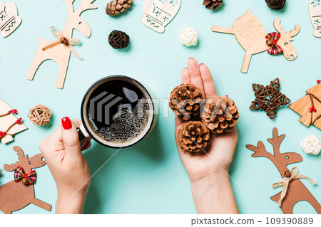 Top view of woman holding a cup of coffee and pine cones in her hands on blue background. Christmas decorations. New year time concept Top view of woman holding a cup of coffee and pine cones in her hands on blue background. Christmas decorations. New year time concept 109390889