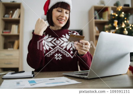 Woman in Santa hat raising fist while looking at laptop with credit card in hand 109391151