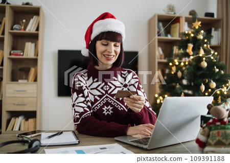 Woman in Santa hat holding blank credit card while typing on computer 109391188