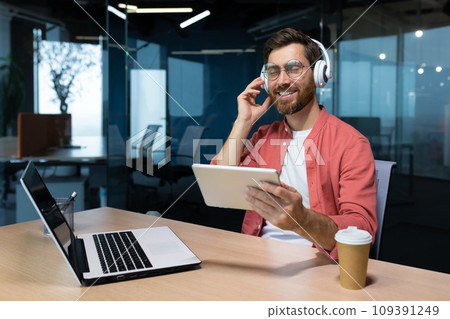 A young freelancer, female student, programmer sits in the office at the table with a laptop, listens to music in headphones, holds tablet in hands. He closed his eyes, rests, relaxes, dances, sings. 109391249