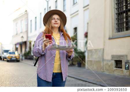 Picture of pretty young woman staying on the street holding phone and map in hands. 30s tourist walking on old city street checks her smartphone. Use technology concept, Traveling Europe in summer 109391650