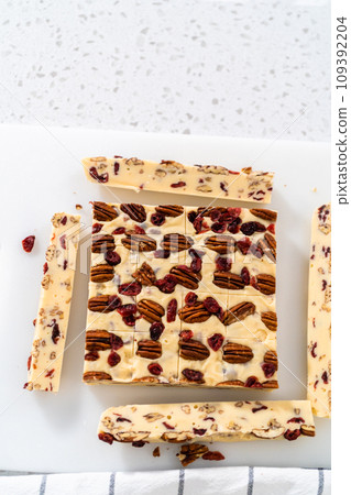 Cutting white chocolate cranberry pecan fudge into small pieces on a white cutting board. 109392204