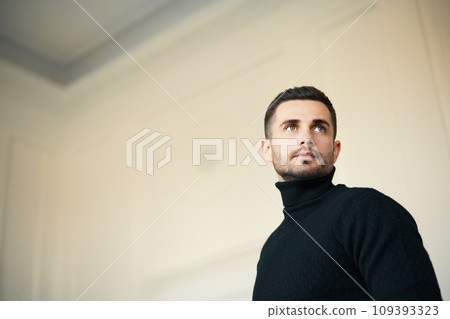Pensive man in a black turtleneck looking upward in a spacious, light-filled room. 109393323