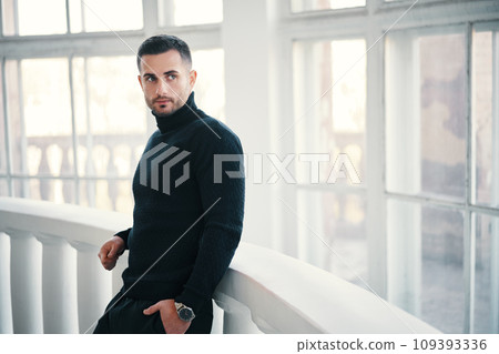 Handsome young man in black turtleneck leans on balcony railings, with large windows in the backgroun 109393336