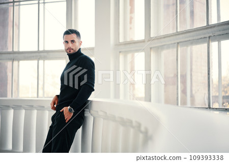 Handsome young man in black turtleneck leans on balcony railings, with large windows in the background Handsome young man in black turtleneck leans on balcony railings, with large windows in the background 109393338