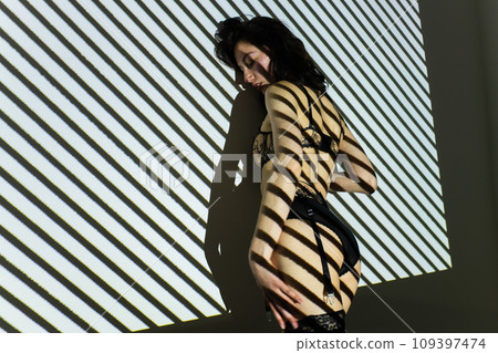 Black and white photo of a beautiful young woman in lace lingerie with a shadow pattern on the face and body in the form of stripes 109397474