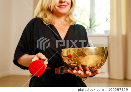 Woman playing on a tibetian singing bowl in cozy room meditating in a yoga Woman playing on a tibetian singing bowl in cozy room meditating in a yoga 109397539