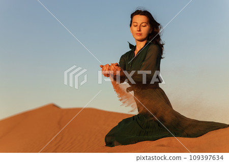 woman in green long dress stands in desert sand dunes background yellow orange sunset 109397634