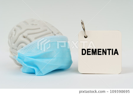 On a white surface there is a brain with a blue mask and a notepad with the inscription - Dementia 109398695