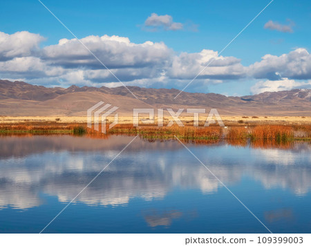 Beautiful autumn mountain lake and mountains. Reflection of snowy peaks and dry yellow grass in the steppe on the surface of a mountain lake. Beautiful autumn mountain lake and mountains. Reflection of snowy peaks and dry yellow grass in the steppe on the surface of a mountain lake. 109399003