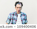 Stupefied man looks with astonishment at camera, being amazed with negative news, does not believe his eyes, wearing checkered shirt and headband. Indoor studio shot isolated on gray background. 109400406