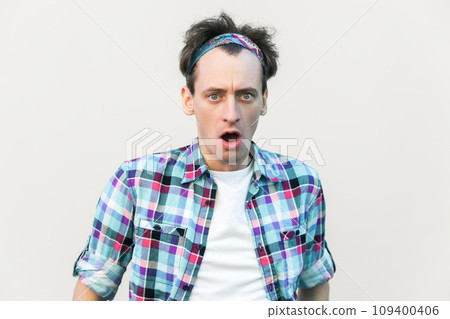 Stupefied man looks with astonishment at camera, being amazed with negative news, does not believe his eyes, wearing checkered shirt and headband. Indoor studio shot isolated on gray background. Stupefied man looks with astonishment at camera, being amazed with negative news, does not believe his eyes, wearing checkered shirt and headband. Indoor studio shot isolated on gray background. 109400406