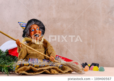 The funny witch Befana and red stocking with sweet coal and candy on beige wall background The funny witch Befana and red stocking with sweet coal and candy on beige wall background 109400567