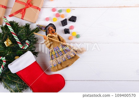 The witch Befana and red stocking with sweet coal and candy on white wooden background 109400570
