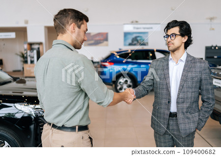 Selective focus of confident car dealer in business suit welcoming young man customer in showroom. Professional salesman shaking client hand in auto dealership. Concept of buying new car. Selective focus of confident car dealer in business suit welcoming young man customer in showroom. Professional salesman shaking client hand in auto dealership. Concept of buying new car. 109400682