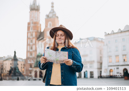 Attractive young female tourist is exploring new city. Redhead girl holding a paper map on Market Square in Krakow. Traveling Europe in autumn. St. Marys Basilica. Vacation concept Attractive young female tourist is exploring new city. Redhead girl holding a paper map on Market Square in Krakow. Traveling Europe in autumn. St. Marys Basilica. Vacation concept 109401513