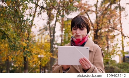 Woman works on tablet walking along town park with yellowed leaves on trees 109402242