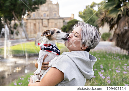Woman with short grey hair holding and kissed on lips by small senior dog in park outdoor. Dog is wearing bandana with an American flag. Joy and companionship between woman and dog jack russell Woman with short grey hair holding and kissed on lips by small senior dog in park outdoor. Dog is wearing bandana with an American flag. Joy and companionship between woman and dog jack russell 109403114