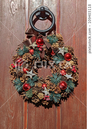 Rustic Christmas wreath hanging on wooden door. Wreath is decorated with natural pine cones, red baubles, and small green fir branches, and few silver stars, dark brown ribbon is tied at the top Rustic Christmas wreath hanging on wooden door. Wreath is decorated with natural pine cones, red baubles, and small green fir branches, and few silver stars, dark brown ribbon is tied at the top 109403118