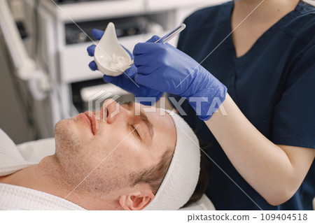 Man laying in cosmetologist cabinet and has a procedure for face skin. Unrecognized female cosmetologist wearing blue medical costume. Woman applying a mask on man's face. Man laying in cosmetologist cabinet and has a procedure for face skin. Unrecognized female cosmetologist wearing blue medical costume. Woman applying a mask on man's face. 109404518